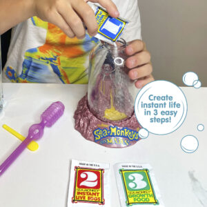 A child wearing a white shirt with bright colors printed on it is holding the Magiquarium with one hand, and pouring powder from a small packet labled with the number 1 with the other hand. A packet with the number 2, a packet with the number 3, a purple pipette, and a yellow plastic spoon all lay on the surface in front of the child. There is a callout in a circle that reads "Create instant life in 3 easy steps!".
